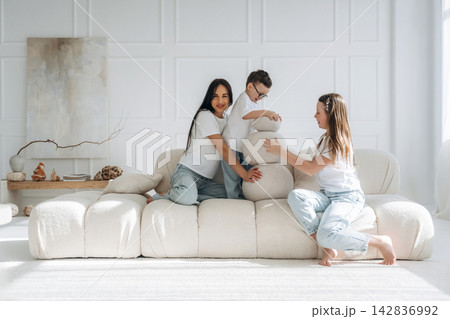 Family of three, two women and a boy, play together on a modern, soft sofa in a bright, minimalist living room with light-colored decor and natural light 142836992