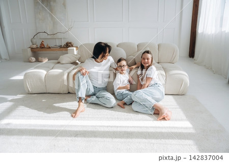 Family of three, including a woman and two children, sitting on a light-colored sofa in a bright, modern living room with large windows and soft furnishings 142837004