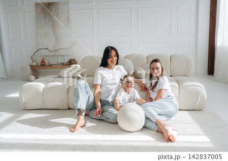 Family of three, including a woman and two girls, sitting on a modern sofa in a bright, minimalist living room with soft furnishings and natural light 142837005