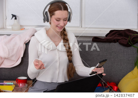 Young girl wearing headphones sits on couch with laptop and phone in hand during an online class at home in the morning Young girl wearing headphones sits on couch with laptop and phone in hand during an online class at home in the morning 142837407