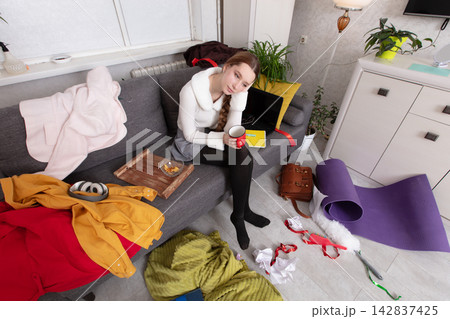 Person sits on couch in messy living room with scattered items and drinks from a mug Person sits on couch in messy living room with scattered items and drinks from a mug 142837425