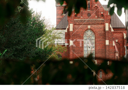 Gothic red brick cathedral facade framed by lush green foliage under soft natural light glare 142837518