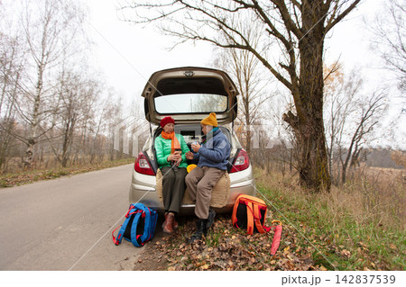 In autumn, an elderly couple enjoys relaxing with warm drinks near a quiet road surrounded by trees and fallen leaves In autumn, an elderly couple enjoys relaxing with warm drinks near a quiet road surrounded by trees and fallen leaves 142837539