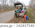 An elderly couple enjoys snacks while sitting by a parked car on the side of the road surrounded by trees in autumn. 142837540