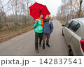 An elderly couple holds a red umbrella while standing on a road in the forest on a cloudy autumn day. 142837541