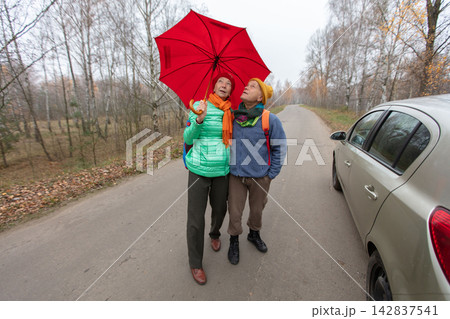 An elderly couple holds a red umbrella while standing on a road in the forest on a cloudy autumn day. 142837541