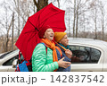 An elderly couple is walking together under a red umbrella on a cloudy day in a forest area wearing warm clothes. 142837542