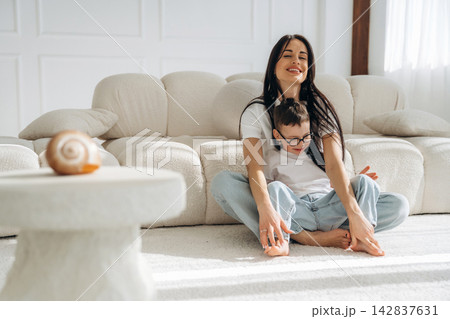 Woman with long dark hair sitting on a light carpeted floor, playing with a young boy in glasses, both smiling in a bright, modern living room with a beige sofa 142837631