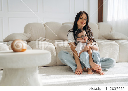 Woman with long dark hair sitting on a light-colored sofa with a child in casual clothing, bright room with natural light and minimalistic decor 142837632