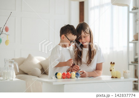 Two children engaged in decorating colorful Easter eggs at a white table in a bright, airy room with soft natural light and minimalistic decor Two children engaged in decorating colorful Easter eggs at a white table in a bright, airy room with soft natural light and minimalistic decor 142837774