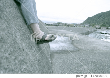 防波堤に腰かけている女性の足元 サンダルを履いている素足の女性の足元の素材写真 防波堤に腰かけている女性の足元 サンダルを履いている素足の女性の足元の素材写真 142838029