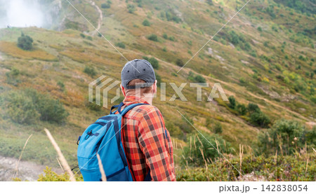 Man with backpack hiking in foggy mountains. Hiker in cap and plaid shirt looking at green valley landscape trail. Active travel, adventure and outdoor recreation concept. Man with backpack hiking in foggy mountains. Hiker in cap and plaid shirt looking at green valley landscape trail. Active travel, adventure and outdoor recreation concept. 142838054