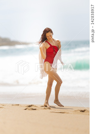 Woman walks on the beach in a red swimsuit while holding a white shirt on a sunny day near the ocean waves Woman walks on the beach in a red swimsuit while holding a white shirt on a sunny day near the ocean waves 142838917
