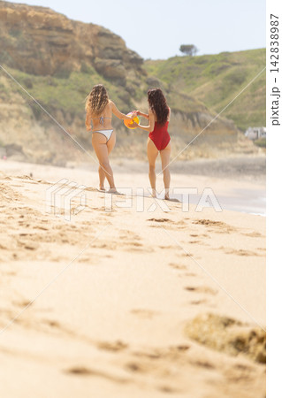 Two women walking on sandy beach holding an orange flotation device, wearing swimsuits, with coastal cliffs and ocean waves in the background under a clear sky 142838987