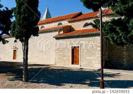 A stone church with red tile roofs and cypress trees stands in Primosten, Croatia. Concept of heritage, religion, architecture, tourism and culture 142839055