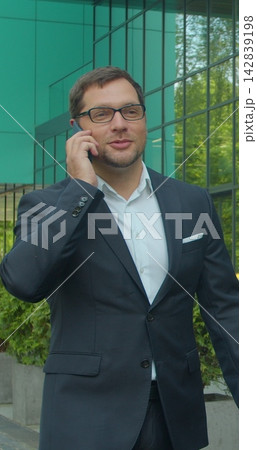 Vertical video. Caucasian businessman in glasses walking the street at the office center and talking on the phone. Close up. Rear. Outside. Vertical video. Caucasian businessman in glasses walking the street at the office center and talking on the phone. Close up. Rear. Outside. 142839198