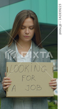 Vertical video. Left to right pan real time portrait shot of businesswoman standing near office center with a sign "looking for job". Vertical video. Left to right pan real time portrait shot of businesswoman standing near office center with a sign "looking for job". 142839223