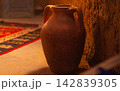 Old clay red pot with two handles standing near wall of samand or plaster against background of blurred background of dark room, illuminated by orange light, on floor carpet with national patterns 142839305