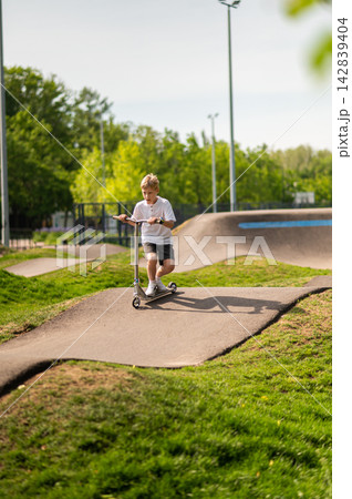 Fair-haired teen riding a scooter in the park on a greenery background Fair-haired teen riding a scooter in the park on a greenery background 142839404