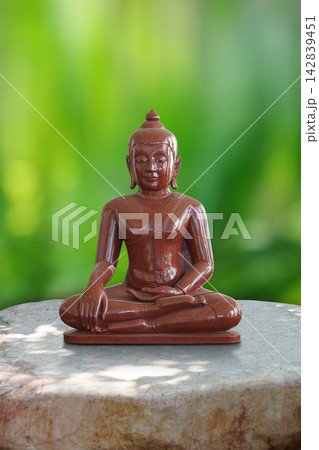 front view brown wooden buddha statue sits in a meditative cross-legged position (Sattvaparyankasana) on stone floor, blur nature green background, religion, buddha, object, decor, copy space front view brown wooden buddha statue sits in a meditative cross-legged position (Sattvaparyankasana) on stone floor, blur nature green background, religion, buddha, object, decor, copy space 142839451