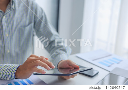 Businessman working in office using tablet while reviewing and analyzing graph data at desk. 142840247