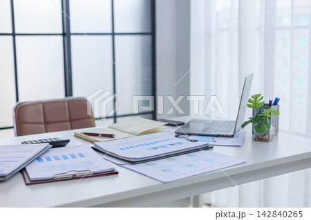 Modern white office workspace of financial or accounting tasks, featuring a laptop, a tablet, financial charts, reports, a calculator, and a notebook on a white desk. 142840265