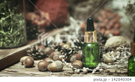 A green glass bottle of natural essential oil among acorns, pinecones and dry herbs on wooden surface, authentic organic extract, selected focus. A green glass bottle of natural essential oil among acorns, pinecones and dry herbs on wooden surface, authentic organic extract, selected focus. 142840304
