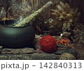 Magic items. Smoking twist of bitter wormwood herb for fumigation in iron cauldron, a ball of red wool, pine bark and dry flowers on wooden background, dark key, selective focus. 142840313