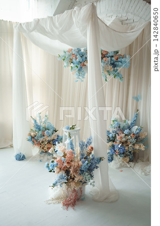 Elegant wedding photo zone with blue and pastel floral decorations, white drapery and wooden stool Elegant wedding photo zone with blue and pastel floral decorations, white drapery and wooden stool 142840650