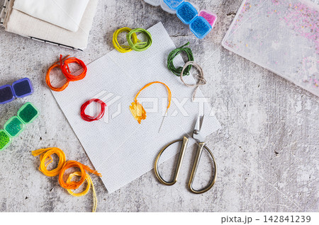 Cross-stitch, Aida cloth, colorful embroidery floss, and scissors on the table top view Cross-stitch, Aida cloth, colorful embroidery floss, and scissors on the table top view 142841239