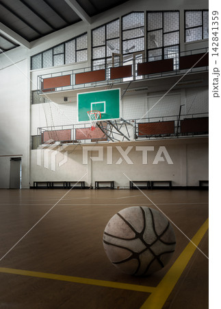 A weathered basketball rests on the court floor in a low-angle perspective, capturing a subtle interplay of light and shadow. Diffused natural light illuminates the ball's worn texture, set against A weathered basketball rests on the court floor in a low-angle perspective, capturing a subtle interplay of light and shadow. Diffused natural light illuminates the ball's worn texture, set against 142841359