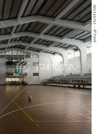 a single basketball sits amidst subtle gradients of light and shadow, framed by soaring structural beams, tiered concrete bleachers, and a vintage green backboard. a single basketball sits amidst subtle gradients of light and shadow, framed by soaring structural beams, tiered concrete bleachers, and a vintage green backboard. 142841386