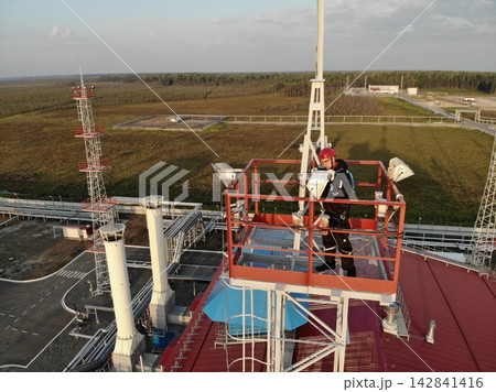 Industrial worker on elevated red platform inspects tower hardware, wearing helmet, gloves, and harness at remote plant. Worker appears concentrated while standing above red roof and utility pipes. Industrial worker on elevated red platform inspects tower hardware, wearing helmet, gloves, and harness at remote plant. Worker appears concentrated while standing above red roof and utility pipes. 142841416