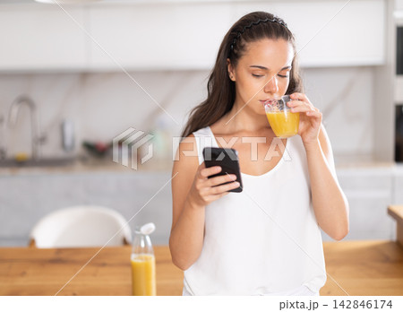 Young woman drinking juice and looking at smartphone in kitchen Young woman drinking juice and looking at smartphone in kitchen 142846174