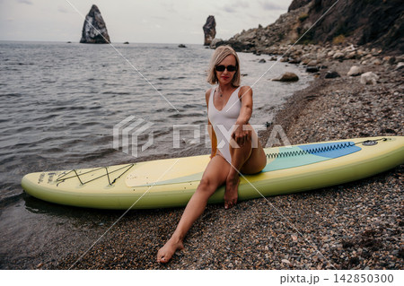 Paddleboard vacation, mature blonde woman posing on yellow board along rocky seashore during overcast day Paddleboard vacation, mature blonde woman posing on yellow board along rocky seashore during overcast day 142850300