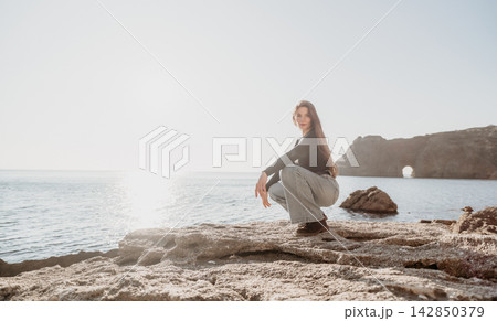 Woman ocean rock crouching on a rocky coastline overlooking the sea with a natural stone arch in the bright background Woman ocean rock crouching on a rocky coastline overlooking the sea with a natural stone arch in the bright background 142850379