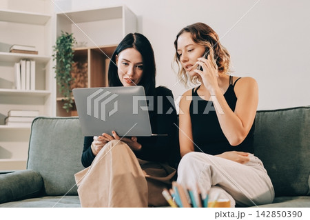 Two young female students using a laptop while sitting on a sofa at home. Student life concept with studying, teamwork, and online learning. Two young female students using a laptop while sitting on a sofa at home. Student life concept with studying, teamwork, and online learning. 142850390