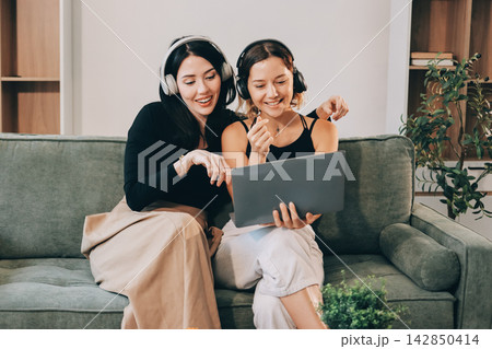 Two young female students using a laptop while sitting on a sofa at home. Student life concept with studying, teamwork, and online learning. 142850414