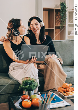 Two young female students using a laptop while sitting on a sofa at home. Student life concept with studying, teamwork, and online learning. Two young female students using a laptop while sitting on a sofa at home. Student life concept with studying, teamwork, and online learning. 142850415