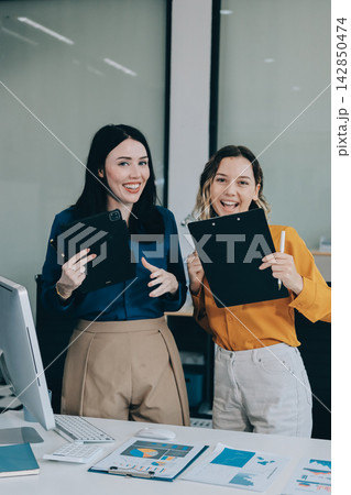 Two businesswomen discussing work using a laptop in a modern office lounge area, showing teamwork and communication 142850474