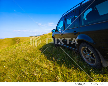 Dark Station Wagon on Grassy Hillside Under Blue Sky Dark Station Wagon on Grassy Hillside Under Blue Sky 142851368
