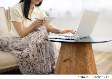 窓辺のソファでノートパソコンを操作しながらノートにメモをする若い女性の手元のクローズアップ 142853712