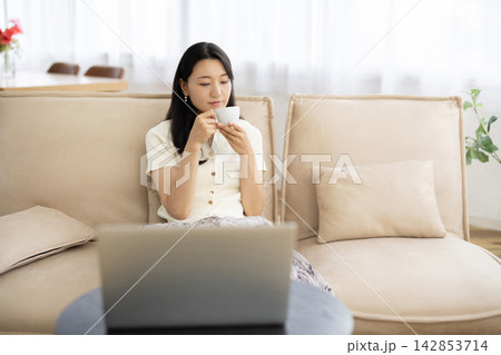 窓辺のソファでノートパソコンを見ながらコーヒーカップを手にリラックスする若い日本人女性 窓辺のソファでノートパソコンを見ながらコーヒーカップを手にリラックスする若い日本人女性 142853714