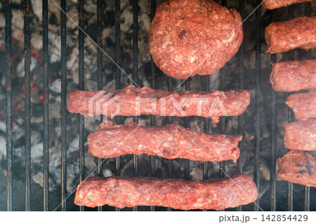 Meat patties and sausage rolls cooking on grill over charcoal. Smoky BBQ scene 142854429