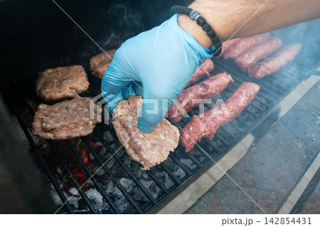 Raw meat patty placed on hot grill with gloved hand. Outdoor BBQ cooking Raw meat patty placed on hot grill with gloved hand. Outdoor BBQ cooking 142854431