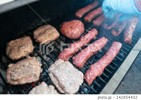 Assorted raw meat patties and sausages grilling over hot coals Assorted raw meat patties and sausages grilling over hot coals 142854432