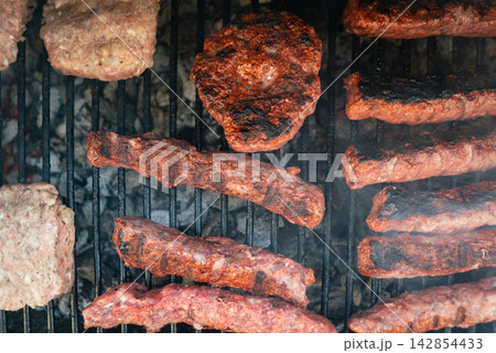 Meat patties and sausage rolls cooking on grill over charcoal. Smoky BBQ scene Meat patties and sausage rolls cooking on grill over charcoal. Smoky BBQ scene 142854433