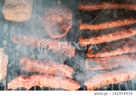 Meat patties and sausage rolls cooking on grill over charcoal. Smoky BBQ scene 142854434