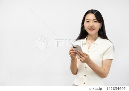 白い壁の前でスマートフォンを持ってカメラ目線で微笑む若い日本人女性 白い壁の前でスマートフォンを持ってカメラ目線で微笑む若い日本人女性 142854605