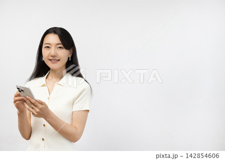 白い壁の前でスマートフォンを持ってカメラ目線で微笑む若い日本人女性 白い壁の前でスマートフォンを持ってカメラ目線で微笑む若い日本人女性 142854606
